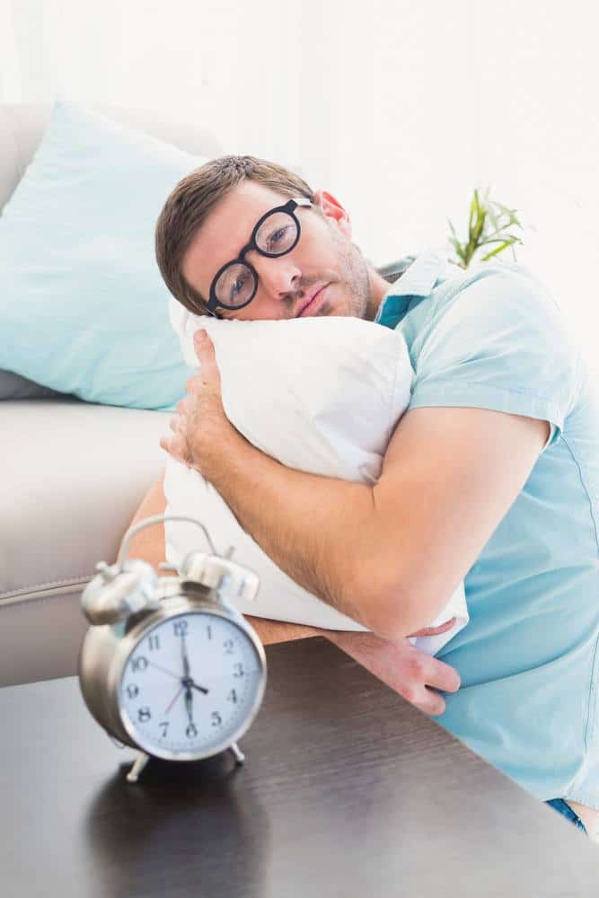 Bored man beside alarm clock hugging a pillow at home in the living room